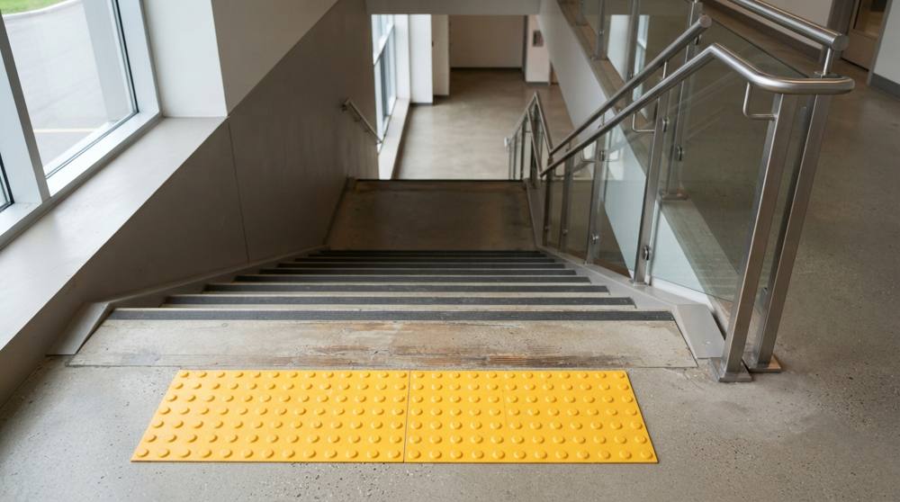 Bande d'éveil de vigilance jaune à plots posée au sol devant la première marche d'un escalier, avec nez de marche contrastés et main courante en inox