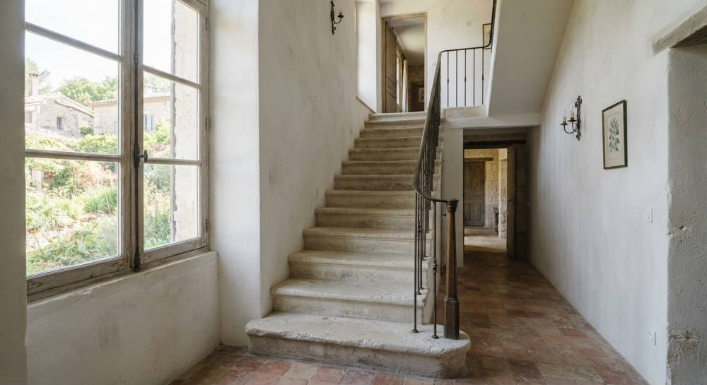 Escalier droit aux marches en pierre calcaire claire, avec une rampe en fer forgé, dans une maison ancienne aux murs chaulés et au sol en terre cuite