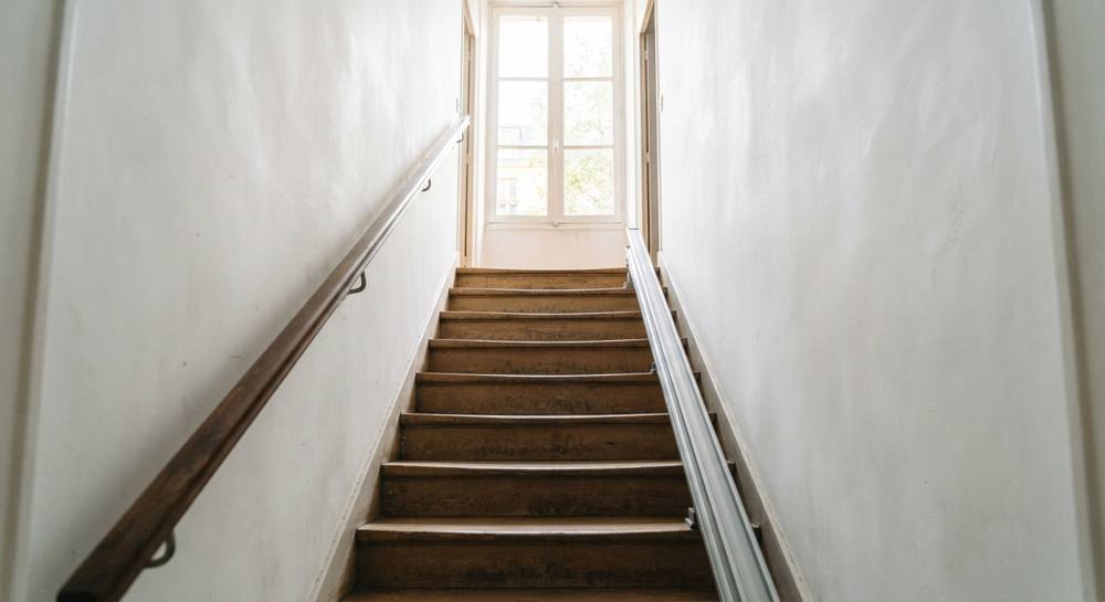 Vue vers le haut d'un escalier droit étroit entre deux murs blancs, avec une main courante en bois à gauche et un rail de monte-escalier le long des marches à droite